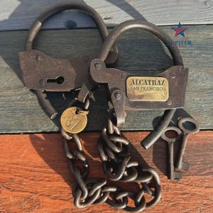 Puede incluir: Grilletes metálicos antiguos con cadena y dos llaves. Los grilletes tienen una placa de latón que dice "ALCATRAZ SAN FRANCISCO". Una etiqueta redonda de latón está adjunta con el número "87".