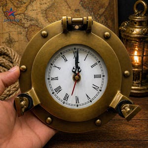 May include: A brass-toned, circular clock designed to look like a ship's porthole. The clock face is white with black Roman numerals and a red second hand. The clock features a hinged top and two side latches, suggesting a nautical style.
