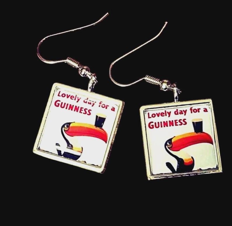 May include: A pair of square earrings with a silver-tone metal frame. Each earring features a white background with a toucan illustration and the text "Lovely day for a GUINNESS". The toucan has a red beak and is holding a pint of Guinness.