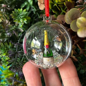 May include: A clear spherical Christmas ornament containing a miniature green champagne bottle with a yellow label reading Moët & Chandon. The bottle is surrounded by small clear beads inside the globe, which hangs from a red ribbon loop.