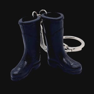 Puede incluir: Un llavero con dos botas de agua en miniatura de color negro unidas a una anilla de metal plateado. Las botas están moldeadas con un acabado oscuro y liso, con detalles realistas que incluyen la suela y la caña, conectadas por una pequeña cadena.