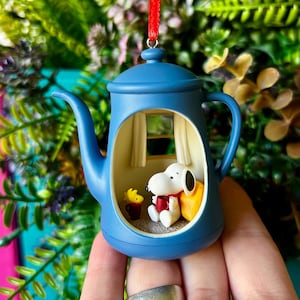 May include: A blue teapot-shaped ornament featuring a cutout window revealing a miniature scene inside. The scene includes a white Snoopy figure holding a red mug, a small yellow Woodstock figure, and a yellow cushion against a window backdrop.