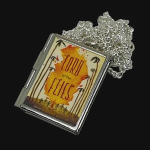 May include: A silver-toned rectangular locket pendant on a matching chain. The front features a miniature book cover illustration for Lord of the Flies, displaying a yellow and orange abstract shape, palm trees, and the title text in dark red.