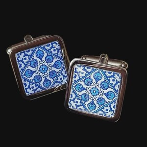 Peut inclure: Une paire de boutons de manchette carrés avec un cadre en métal argenté. Le centre de chaque bouton de manchette présente un motif floral bleu et blanc. Les boutons de manchette sont posés sur un fond sombre.