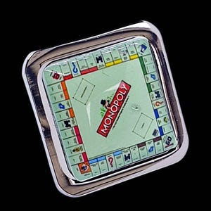 May include: A square-shaped pin featuring a miniature Monopoly game board graphic under a clear, glossy dome. The board displays the iconic red Monopoly logo, colorful property spaces, and game board markings set within a polished silver-tone metal frame.