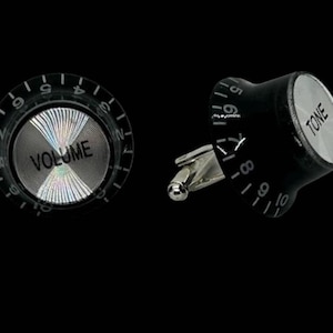 May include: A pair of cufflinks designed to resemble guitar amplifier knobs. One cufflink features the word "VOLUME" on a silver face, while the other says "TONE". Both are black with white number markings.