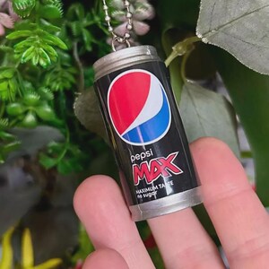 May include: Miniature Pepsi Max can keyring. The black can features the Pepsi logo in red, white, and blue. Text reads "pepsi MAX" and "MAXIMUM TASTE no sugar". The keyring has a silver chain and is a small replica of the drink.