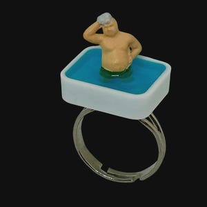 May include: A ring featuring a rectangular white basin filled with translucent blue resin. A small, light-skinned figure with gray hair is submerged in the blue material, with one arm raised to the head and the other resting on the hip.