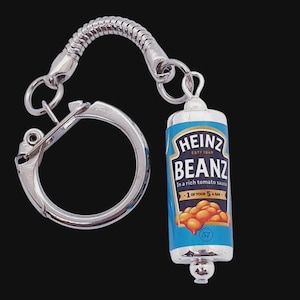 May include: A silver-coloured metal keyring featuring a miniature cylindrical charm shaped like a Heinz Beanz tin. The charm displays the blue Heinz Beanz label with the text In a rich tomato sauce and 1 of your 5 a day, plus a graphic of baked beans.