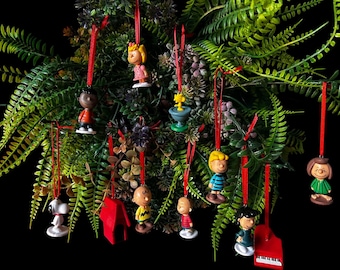 Może przedstawiać: Kolekcja ozdób przedstawiających postacie z serii Peanuts zawieszonych na czerwonych wstążkach, w tym Snoopy, Charlie Brown, Lucy, Linus, Sally, Franklin i Schroeder z pianinem, a także czerwona buda dla psa i poidełko dla ptaków z kolorowymi detalami.