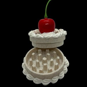 May include: A round, beige, speckled herb grinder designed to look like a small cake. The lid features a glossy red cherry with a green stem on top. The base and lid are decorated with white piped frosting details, and the interior contains teeth.