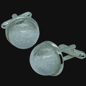 May include: A pair of silver cufflinks with a clear, spherical design. Each cufflink features a glass-like orb filled with a textured, white material. The cufflinks have a classic, T-bar closure and are set against a black background.