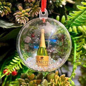 May include: A clear, round Christmas ornament with a miniature Prosecco bottle inside. The bottle has a gold foil top and a label that reads "ROSECC". The ornament is filled with clear, iridescent beads and hangs from a red ribbon.