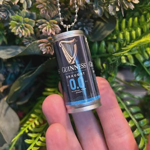 May include: Miniature Guinness Draught 0.0% stout can pendant. The black and blue can features the Guinness logo and text. The pendant hangs from a silver ball chain. The background is blurred green foliage.