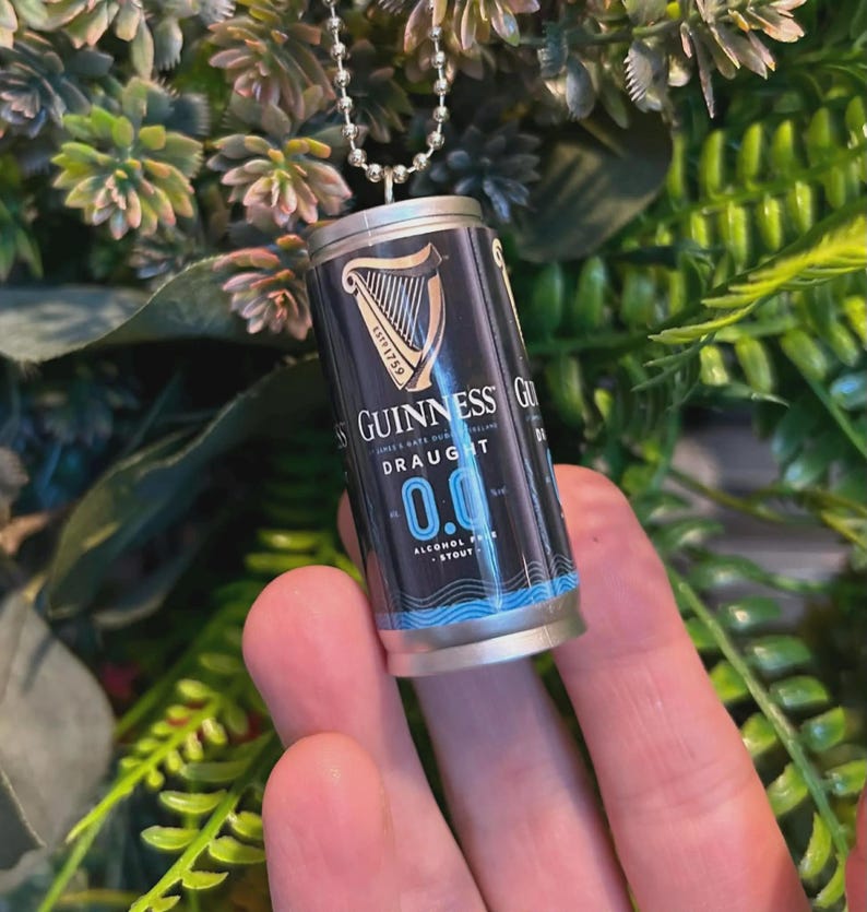 May include: Miniature Guinness Draught 0.0% stout can pendant. The black and blue can features the Guinness logo and text. The pendant hangs from a silver ball chain. The background is blurred green foliage.
