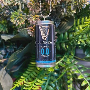 May include: A miniature Guinness Draught 0.0 can pendant, suspended by a silver ball chain. The can features the Guinness logo and branding, with a black and blue color scheme. The background is a mix of green foliage.