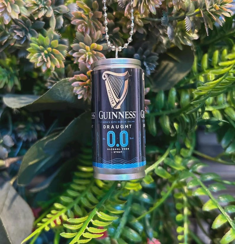 May include: A miniature Guinness Draught 0.0 can pendant, suspended by a silver ball chain. The can features the Guinness logo and branding, with a black and blue color scheme. The background is a mix of green foliage.