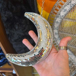 May include: A crescent moon decoration covered in small, reflective mirrored tiles. The silver-colored moon hangs from a delicate chain. The decorative piece is held in a hand.