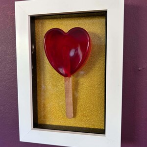 May include: A framed wall art piece featuring a translucent red heart-shaped lollipop with a wooden stick. The object is mounted against a textured gold glitter background and enclosed within a simple white rectangular frame with a black inner border.