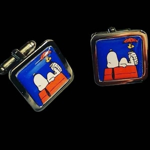 以下が含まれることがあります： シルバーカラーのメタルフレームが付いた、正方形のカフスボタンのペア。各カフスボタンは、スヌーピーが犬小屋に横たわっている漫画の画像が描かれた青色の背景が特徴です。犬小屋はオレンジ色で、スヌーピーは白です。
