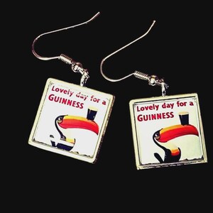 May include: A pair of square earrings with a silver-tone metal frame. Each earring features a white background with a toucan illustration and the text "Lovely day for a GUINNESS". The toucan has a red beak and is holding a pint of Guinness.