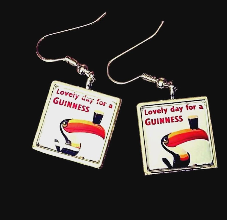 May include: A pair of square earrings with a silver-tone metal frame. Each earring features a white background with a toucan illustration and the text "Lovely day for a GUINNESS". The toucan has a red beak and is holding a pint of Guinness.