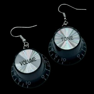 May include: A pair of drop earrings featuring black circular knobs with silver-toned metallic tops. One knob is labelled VOLUME and the other TONE, both with black numerical markings from zero to ten and silver wire hooks for attachment.