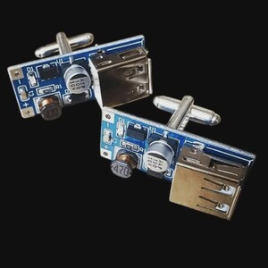 Op de afbeelding: Een paar unieke manchetknopen met een blauw printplaatontwerp en zilverkleurige hardware. Elke manchetknop heeft een USB-poort en een zilverkleurige tuimelsluiting. De achtergrond is zwart.
