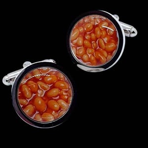 May include: A pair of novelty cufflinks featuring a close-up image of baked beans in a tomato sauce. The round, silver-toned cufflinks have a black border around the bean image. The background is black.
