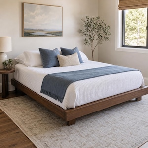 May include: A low-profile wooden platform bed frame with a white textured quilt, two blue square pillows, one small cream-colored rectangular pillow, and a blue throw blanket with fringe draped across the foot of the bed.