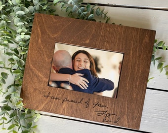 Op de afbeelding: Een bruin houten fotolijstje met een foto van twee mensen die elkaar omhelzen. Het lijstje heeft de woorden "Ik ben trots op je" en een handtekening die "Love Dad" zegt.