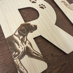 May include: Wooden letter "R" with a detailed engraving of a dog and paw prints. The letter is made of light-colored wood with a dark brown engraved design. Another wooden letter with a bird engraving is partially visible in the background.