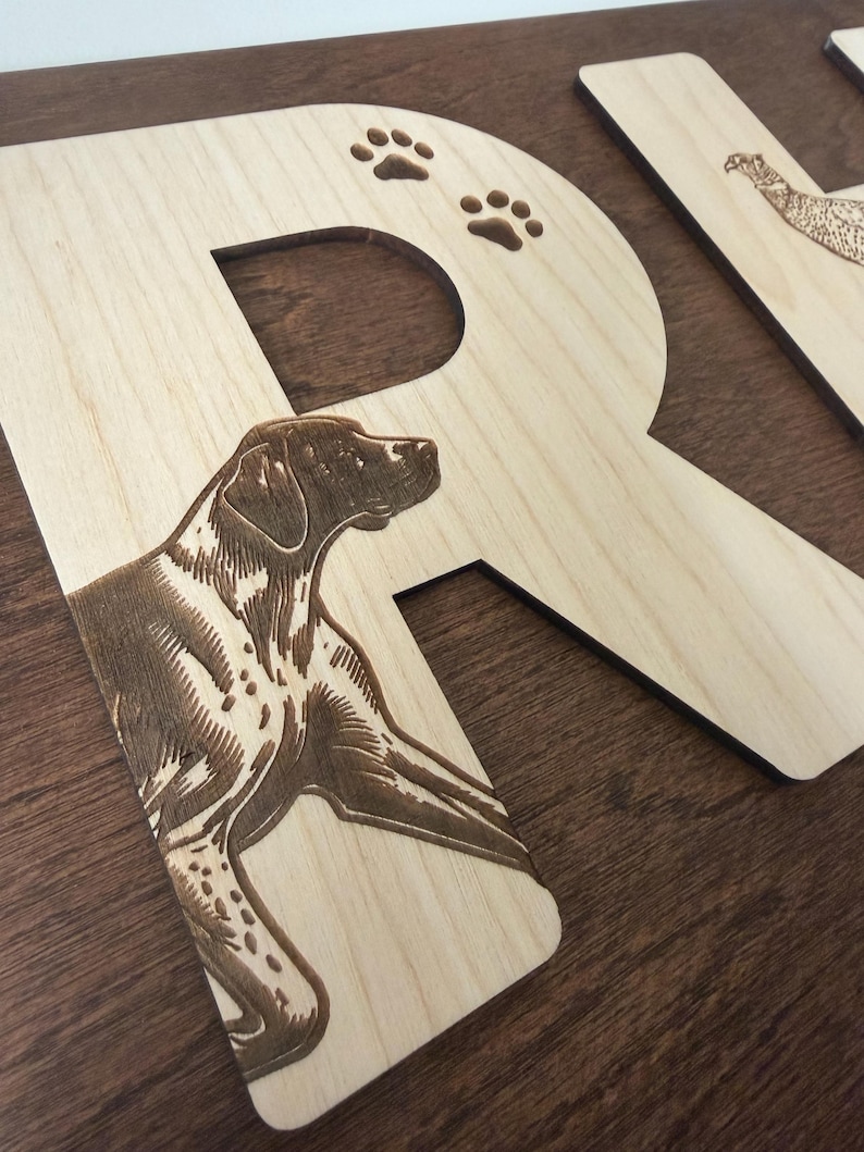 May include: Wooden letter "R" with a detailed engraving of a dog and paw prints. The letter is made of light-colored wood with a dark brown engraved design. Another wooden letter with a bird engraving is partially visible in the background.