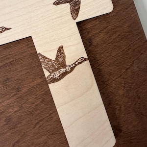 May include: Wooden letter "T" with engraved flying ducks. The ducks are detailed with brown feathers and are in mid-flight. The wood has a light, natural finish, contrasting with the darker brown background.