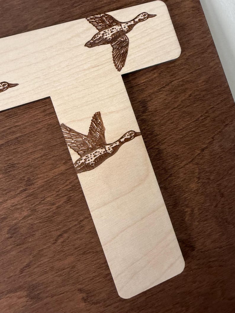 May include: Wooden letter "T" with engraved flying ducks. The ducks are detailed with brown feathers and are in mid-flight. The wood has a light, natural finish, contrasting with the darker brown background.