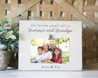 May include: White picture frame with a clip at the top. The frame has a photo of a family of four in a park. The text on the frame reads "Our favorite people call us Grammie and Grandpa Avery & Ella".
