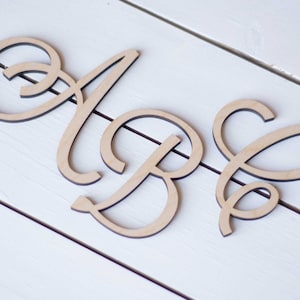 May include: Three wooden letters, A, B, and C, are arranged in a row on a white wooden surface. The letters are in a cursive font and are painted a light brown color.