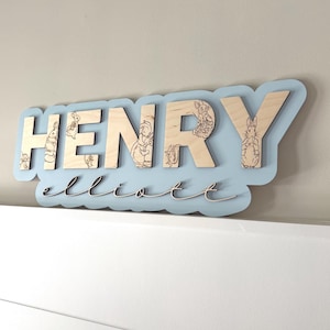 May include: A custom wooden wall sign featuring the name HENRY in large block letters and elliott in script, mounted on a light blue scalloped backing. The block letters are engraved with detailed illustrations of rabbits and small woodland creatures.