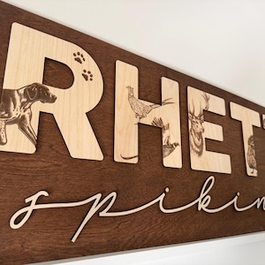 May include: Wooden sign with the name "Rhett" in large, light-colored letters, each featuring a different animal illustration. The name "Spiking" is written in a cursive font below. The background is a dark brown.