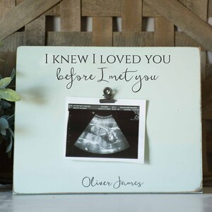 May include: A light blue wooden picture frame with the text "I knew I loved you before I met you" and a black and white ultrasound image. The frame has a clip to hold the ultrasound image and the name "Oliver James" is written below the image.