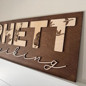 May include: Wooden sign with the name "RHETT" in large, light-colored letters, each featuring a different animal illustration. The name "spiking" is written in cursive below. The sign is mounted on a dark brown wooden background.