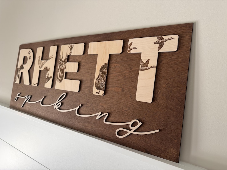 May include: Wooden sign with the name "RHETT" in large, light-colored letters, each featuring a different animal illustration. The name "spiking" is written in cursive below. The sign is mounted on a dark brown wooden background.