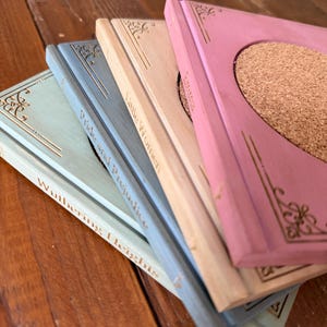May include: A set of four square coasters designed to look like miniature book spines, featuring pastel colors including pink, beige, blue, and light green. Each coaster has a circular cork center and gold-colored embossed book titles.