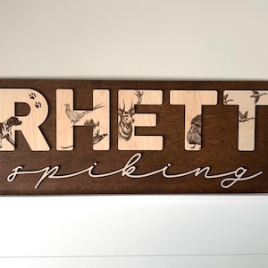 May include: Wooden sign with the name "Rhett" in light wood letters on a dark brown background. The letters feature engraved images of a dog, birds, deer, and ducks. The word "spiking" is written in cursive below.