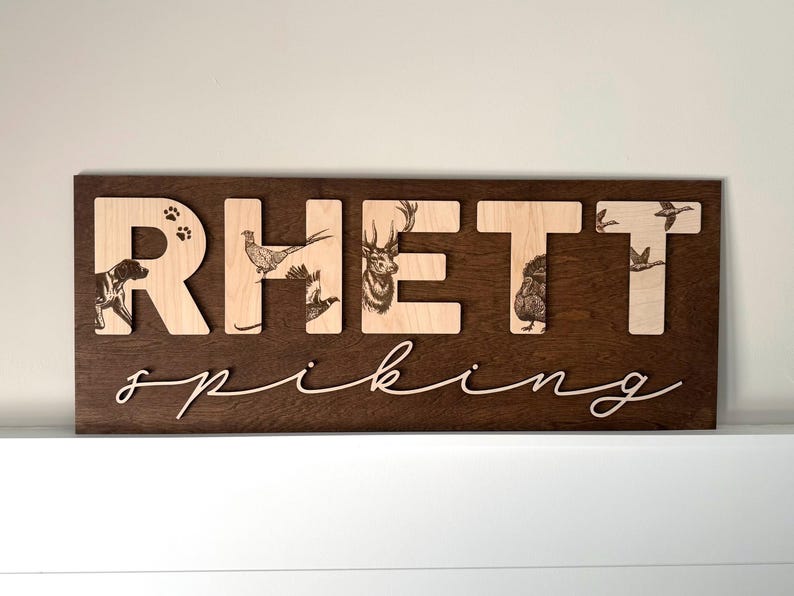 May include: Wooden sign with the name "Rhett" in light wood letters on a dark brown background. The letters feature engraved images of a dog, birds, deer, and ducks. The word "spiking" is written in cursive below.