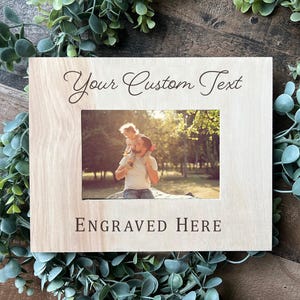 May include: A wooden picture frame with a light brown finish. The frame has a rectangular opening for a photo and a space for engraving. The text "Your Custom Text" is engraved at the top of the frame and the text "Engraved Here" is engraved at the bottom of the frame. The frame is surrounded by green foliage.