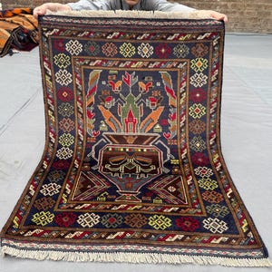 May include: A rectangular, hand-knotted rug with a central design featuring a vase with floral elements in red, orange, and green. The rug has a dark blue background with a border of repeating geometric patterns in red, yellow, and brown. The rug's dimensions are approximately 36 x 48 inches.