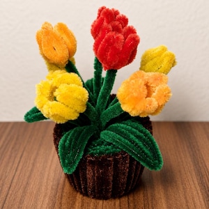 Könnte beinhalten: Ein dekoratives Arrangement aus fünf Blumen, die aus flauschigen Pfeifenreinigern in den Farben Rot, Orange und Gelb gefertigt sind. Die Blumen haben grüne Stiele und Blätter und stecken in einem kleinen, braunen, gerippten Pfeifenreiniger-Topf.