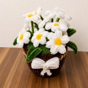 Könnte beinhalten: Ein handgefertigter Strauß aus weißen Pfeifenreiniger-Gänseblümchen mit leuchtend gelben Mitten und grünen Stielen, arrangiert in einem kleinen, dunkelbraunen, strukturierten Topf. Der Topf ist mit einer weißen Pfeifenreiniger-Schleife verziert.