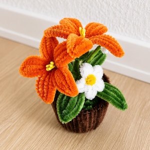 Könnte beinhalten: Ein handgefertigtes Blumenarrangement aus Pfeifenreinigern mit zwei leuchtend orangefarbenen Lilien mit gelben Zentren und einem weißen Gänseblümchen mit gelbem Zentrum. Die Blumen stecken in einer dunkelbraunen, gerippten, becherförmigen Basis mit grünen Blättern.